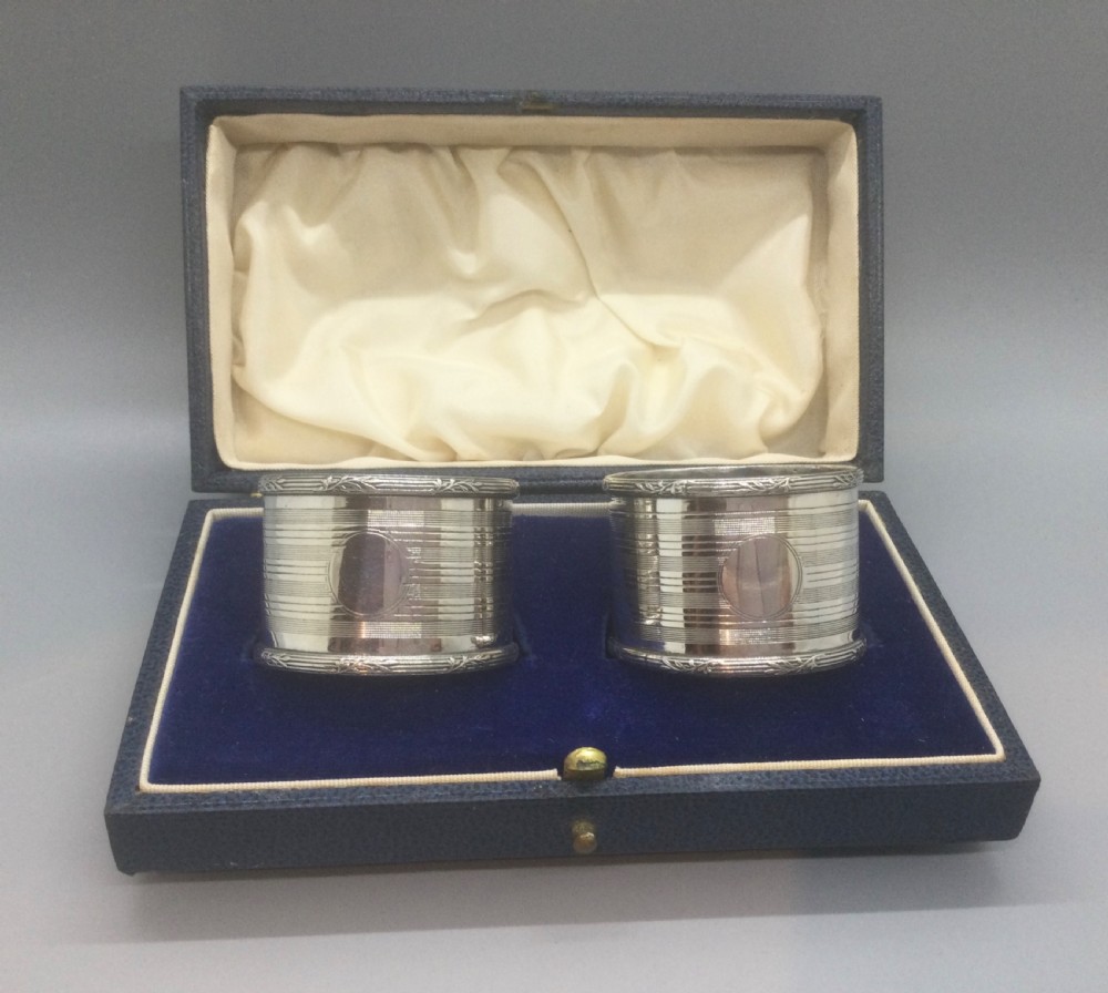 A pair of engine-turned sterling silver napkin rings with vacant oval cartouches, presented inside a blue velvet-lined fitted display case.