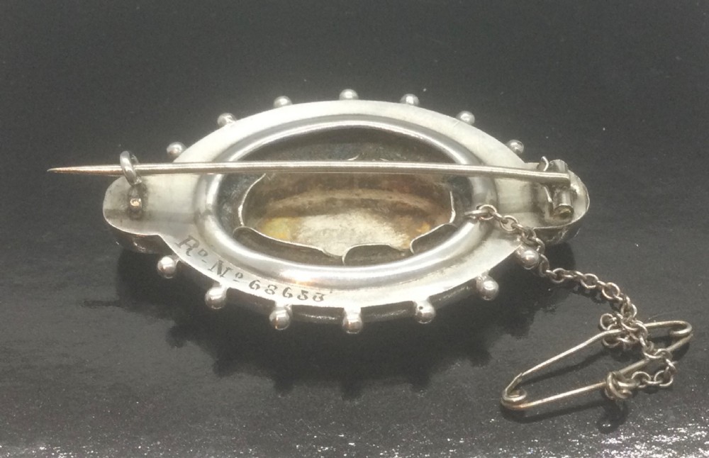 An oval Victorian silver brooch decorated with hand-painted yellow enamel flowers, green leaves, beaded outer edges, and a central silver crest motif. View IV