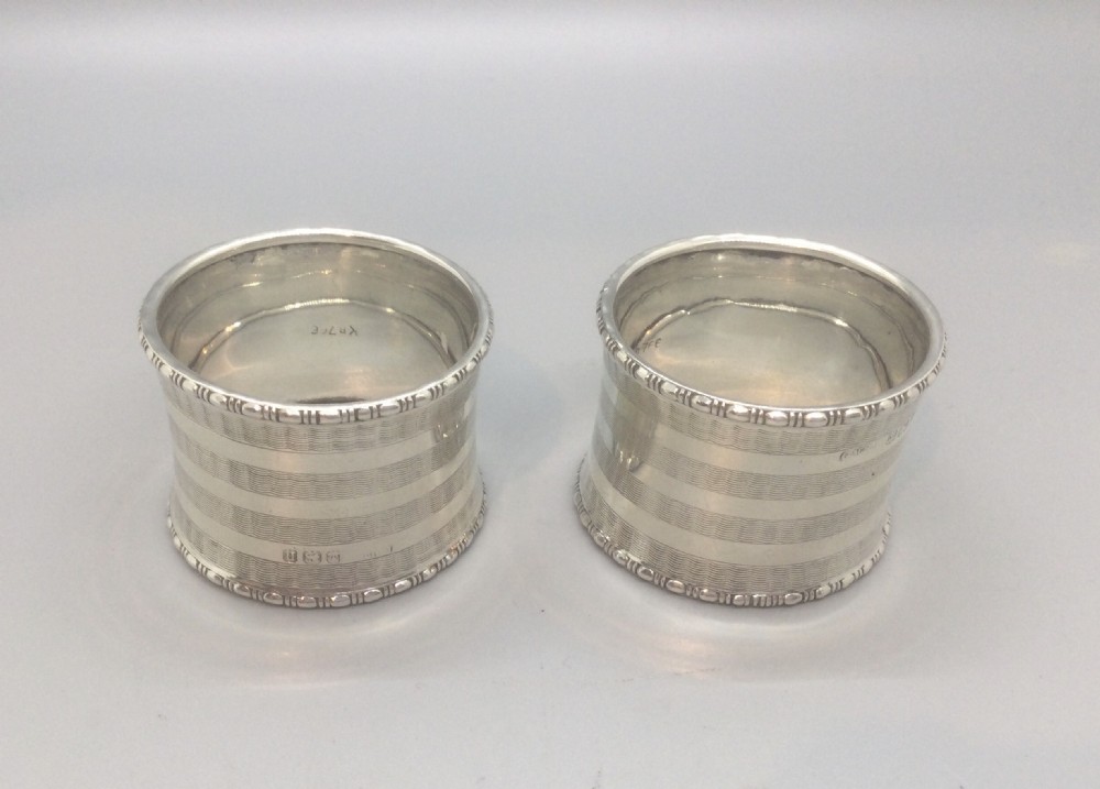 A pair of engine-turned sterling silver napkin rings with blank circular cartouches, displayed inside a black box with blue velvet and silk lining. View IV