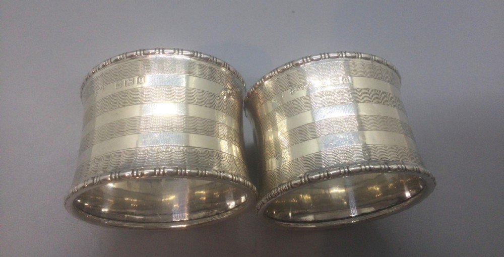 A pair of engine-turned sterling silver napkin rings with blank circular cartouches, displayed inside a black box with blue velvet and silk lining. View V