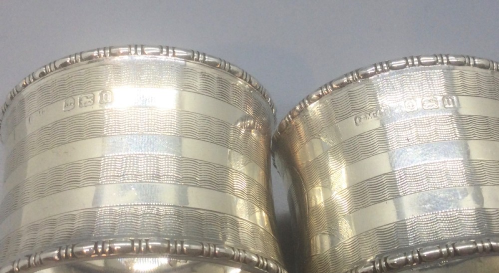 A pair of engine-turned sterling silver napkin rings with blank circular cartouches, displayed inside a black box with blue velvet and silk lining. View VI