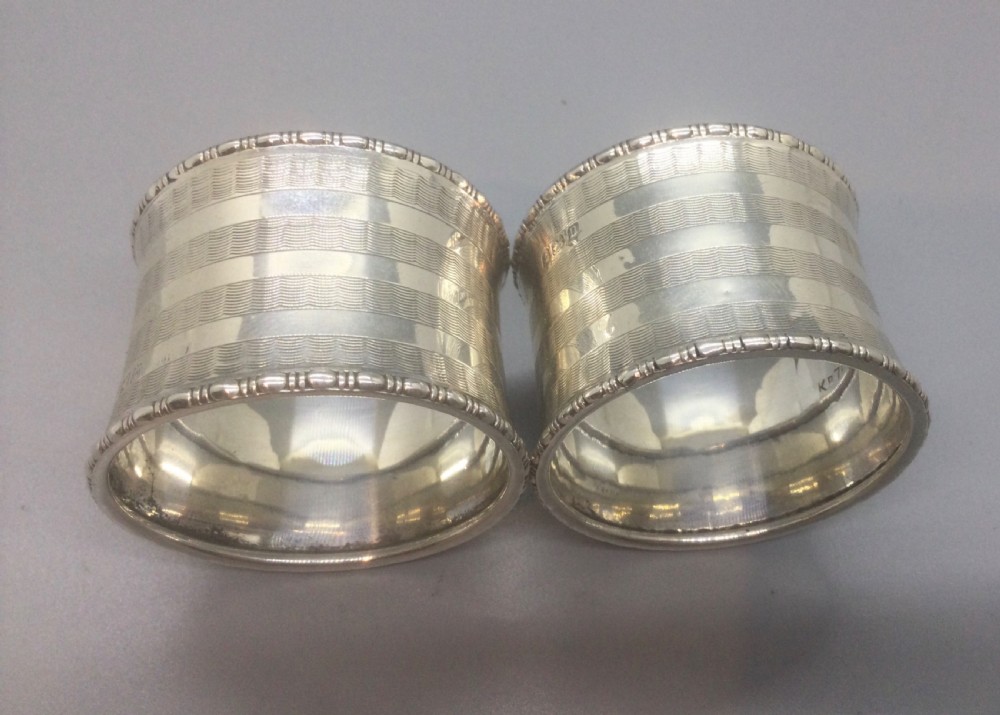 A pair of engine-turned sterling silver napkin rings with blank circular cartouches, displayed inside a black box with blue velvet and silk lining. View VII