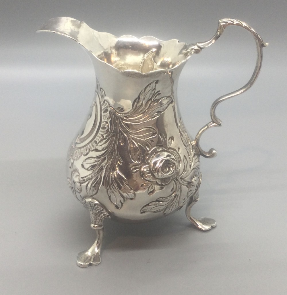 A George II sterling silver cream jug with hand-chased floral repousse decoration, a scalloped rim, scroll handle, and three pad feet.