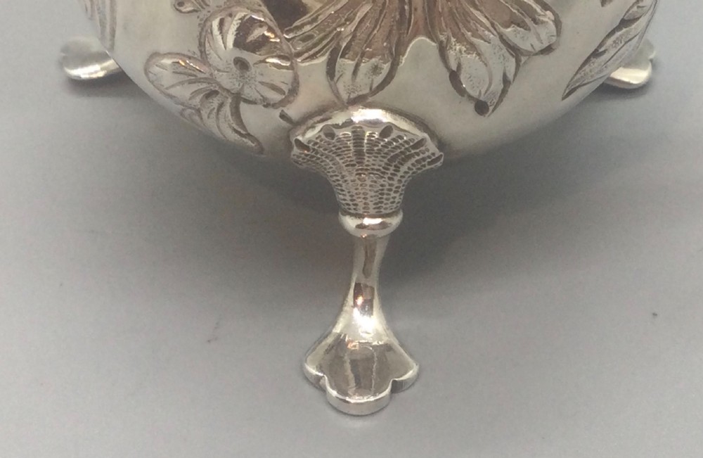 A George II sterling silver cream jug with hand-chased floral repousse decoration, a scalloped rim, scroll handle, and three pad feet. View IV