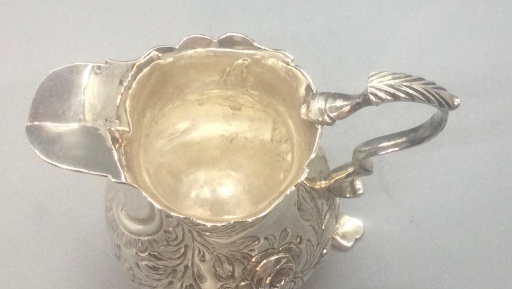 A George II sterling silver cream jug with hand-chased floral repousse decoration, a scalloped rim, scroll handle, and three pad feet. View VII