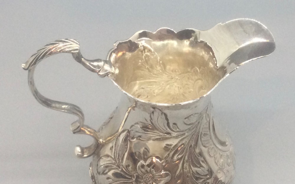 A George II sterling silver cream jug with hand-chased floral repousse decoration, a scalloped rim, scroll handle, and three pad feet. View VIII