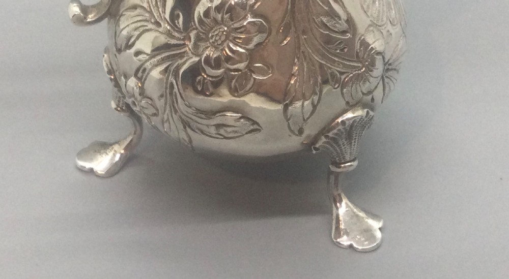 A George II sterling silver cream jug with hand-chased floral repousse decoration, a scalloped rim, scroll handle, and three pad feet. View IX