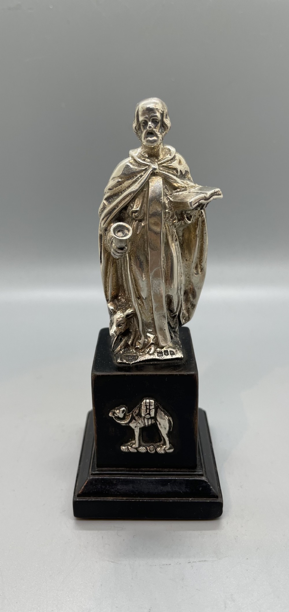 An Edwardian sterling silver figure of an apostle holding a chalice and open book, mounted on a dark square wooden plinth featuring a silver camel motif.