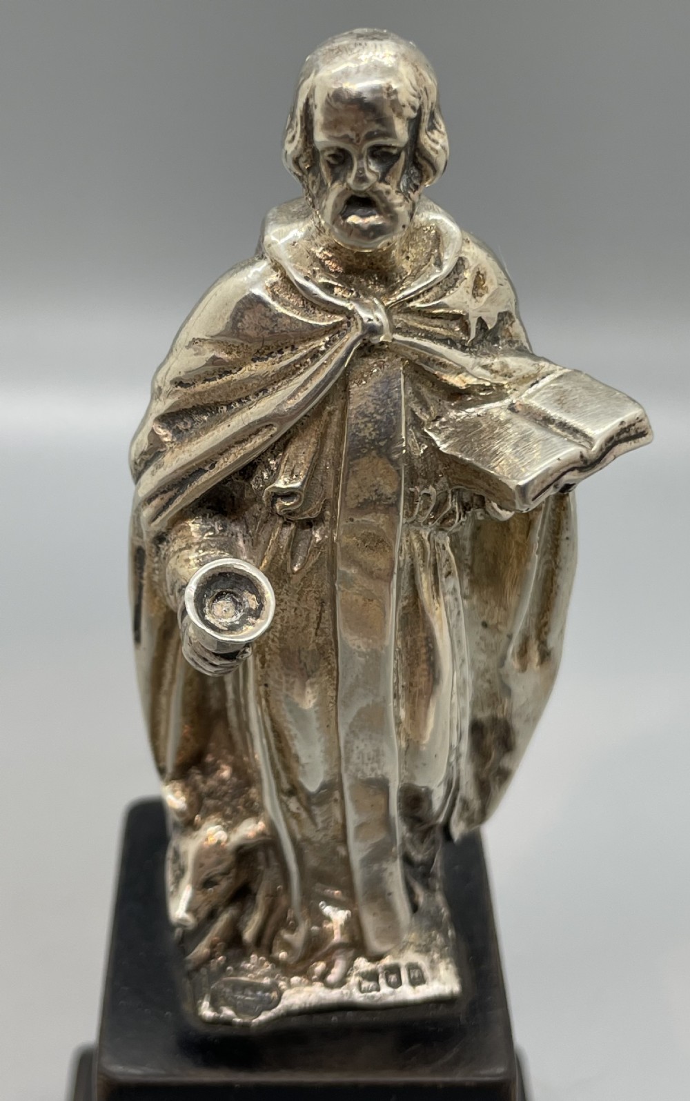 An Edwardian sterling silver figure of an apostle holding a chalice and open book, mounted on a dark square wooden plinth featuring a silver camel motif. View VI
