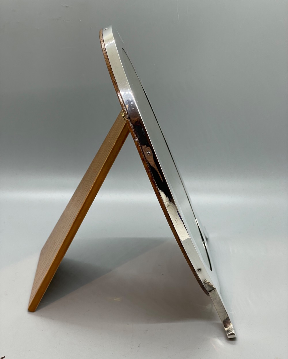 An oval sterling silver photo frame featuring two outward-curving bracket feet and a wooden easel back support. View VI
