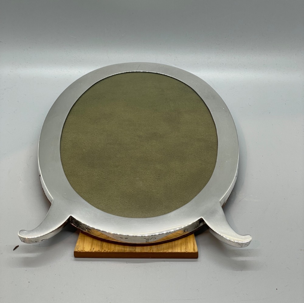 An oval sterling silver photo frame featuring two outward-curving bracket feet and a wooden easel back support. View X