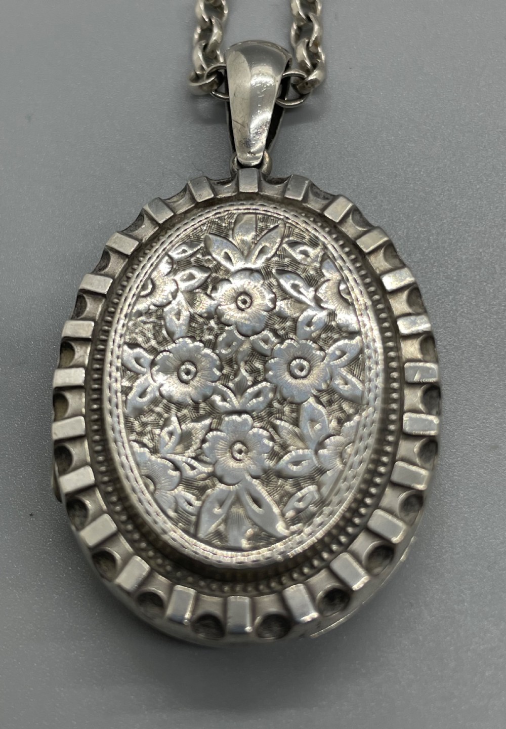 An oval Victorian silver pendant locket decorated with floral engravings and a crenellated outer rim suspended from a silver chain. View IV