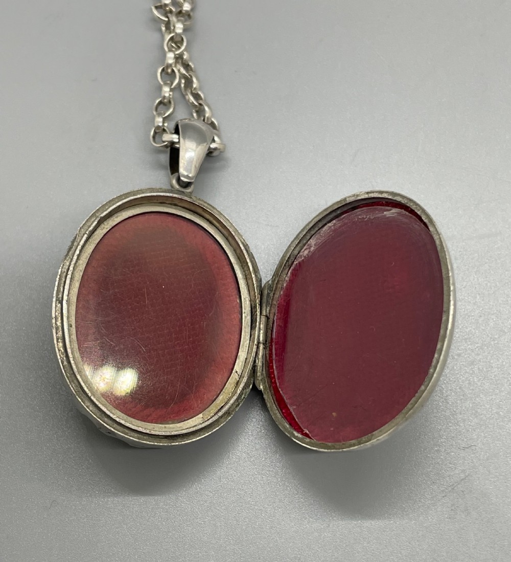 An oval Victorian silver pendant locket decorated with floral engravings and a crenellated outer rim suspended from a silver chain. View VI