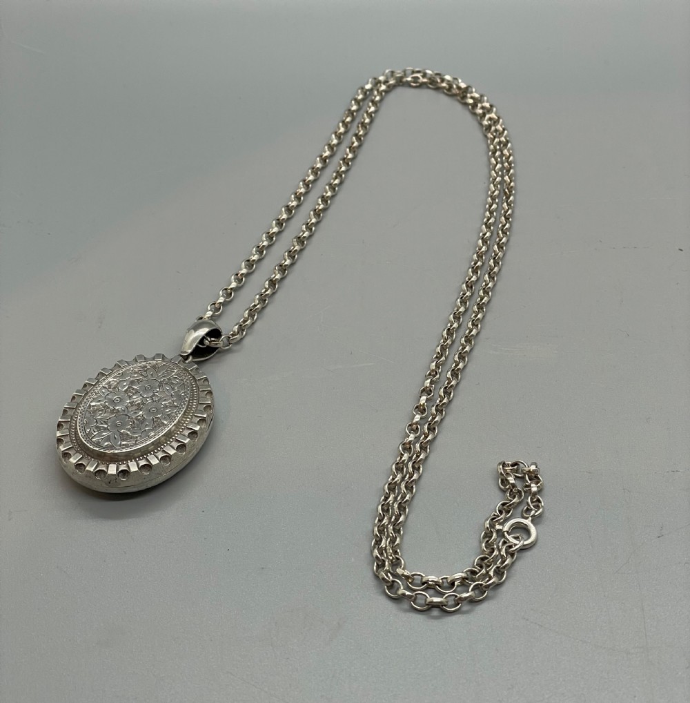 An oval Victorian silver pendant locket decorated with floral engravings and a crenellated outer rim suspended from a silver chain. View VII