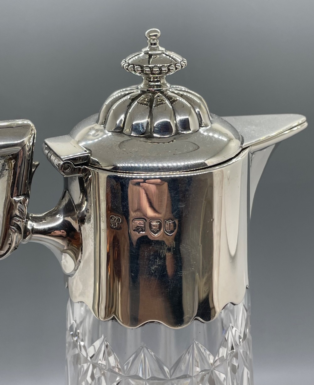 A late Victorian cut-crystal claret jug featuring a fluted glass body, hallmarked sterling silver mounts, a hinged lid, and an angular handle. View V