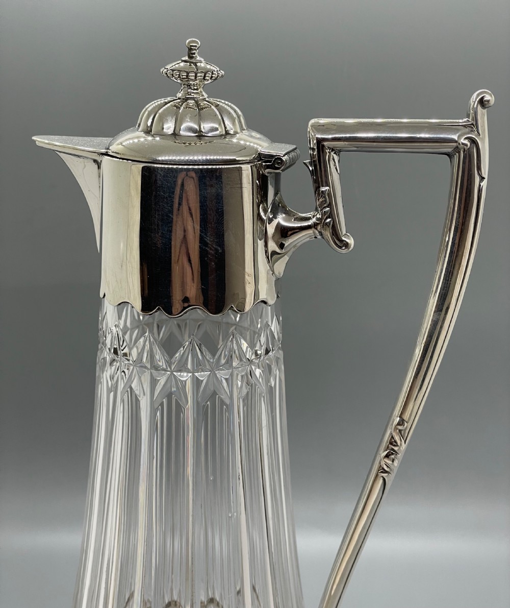 A late Victorian cut-crystal claret jug featuring a fluted glass body, hallmarked sterling silver mounts, a hinged lid, and an angular handle. View X