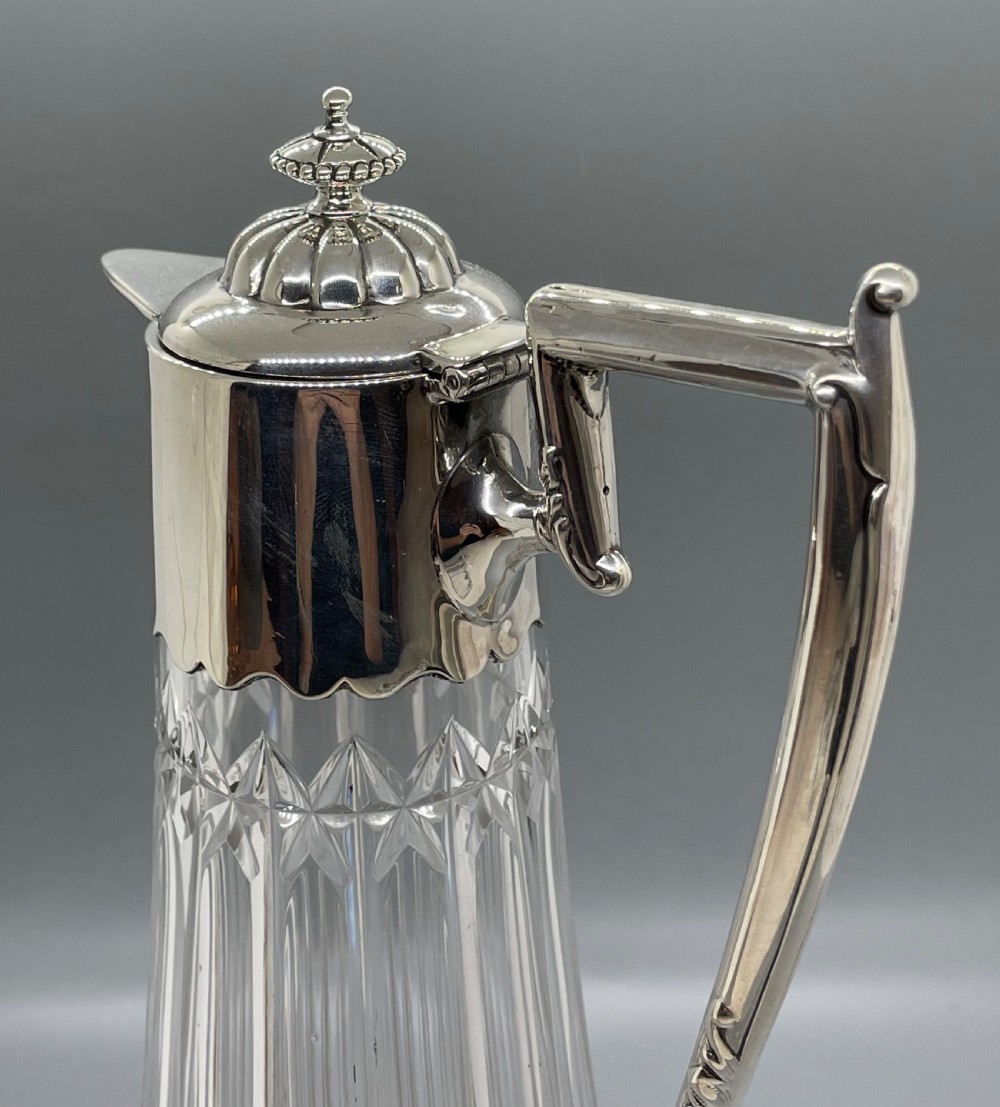 A late Victorian cut-crystal claret jug featuring a fluted glass body, hallmarked sterling silver mounts, a hinged lid, and an angular handle. View XIII