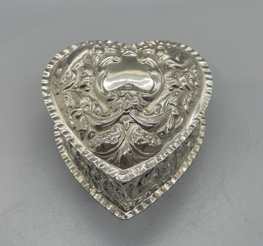 A heart-shaped sterling silver trinket box decorated with relief foliate scrollwork, a crimped rim, and a smooth central cartouche.