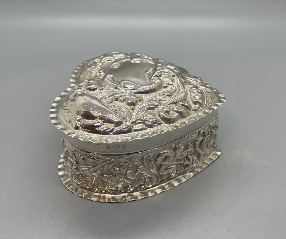 A heart-shaped sterling silver trinket box decorated with relief foliate scrollwork, a crimped rim, and a smooth central cartouche. View IV