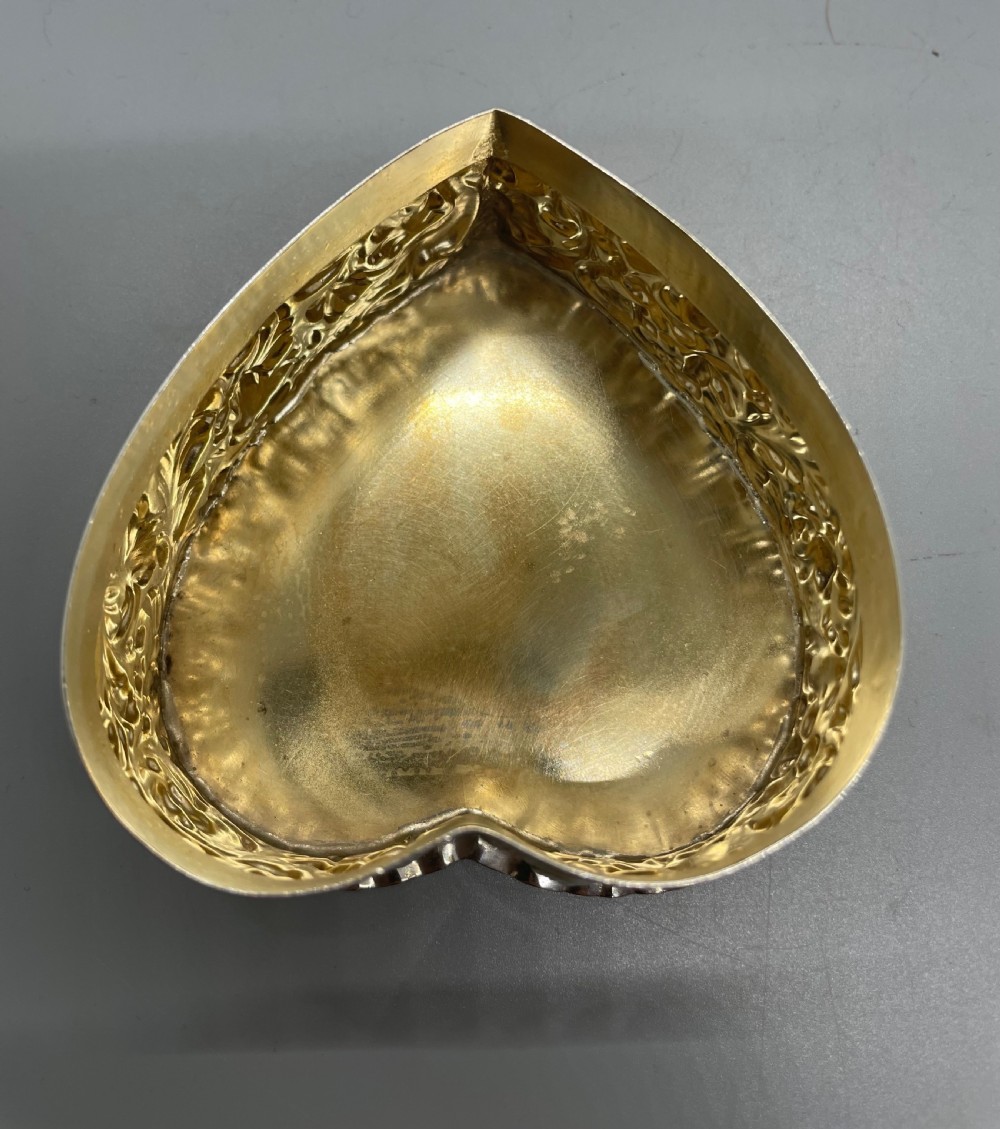 A heart-shaped sterling silver trinket box decorated with relief foliate scrollwork, a crimped rim, and a smooth central cartouche. View X