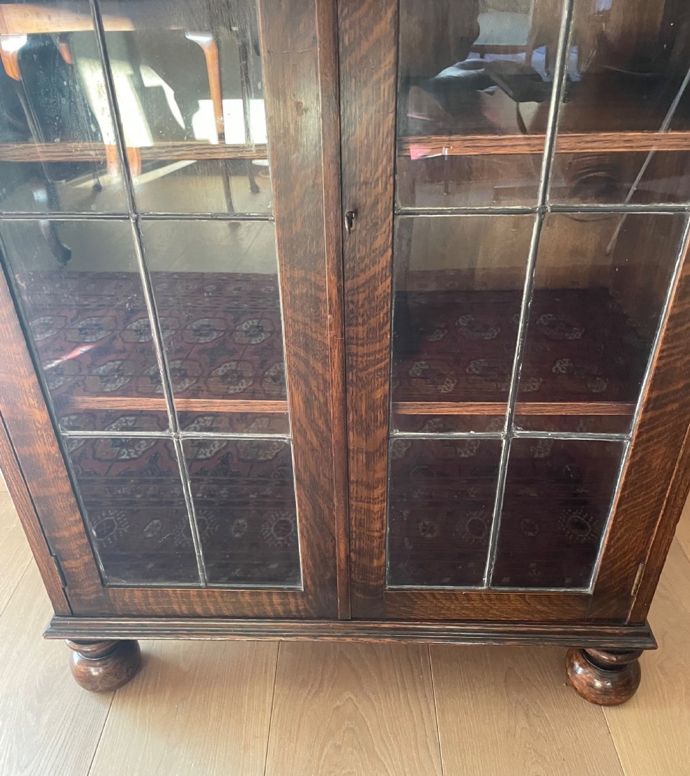 A 1920s dark oak bookcase featuring twin glazed doors with leaded glass panes, set on turned bun feet. View VII