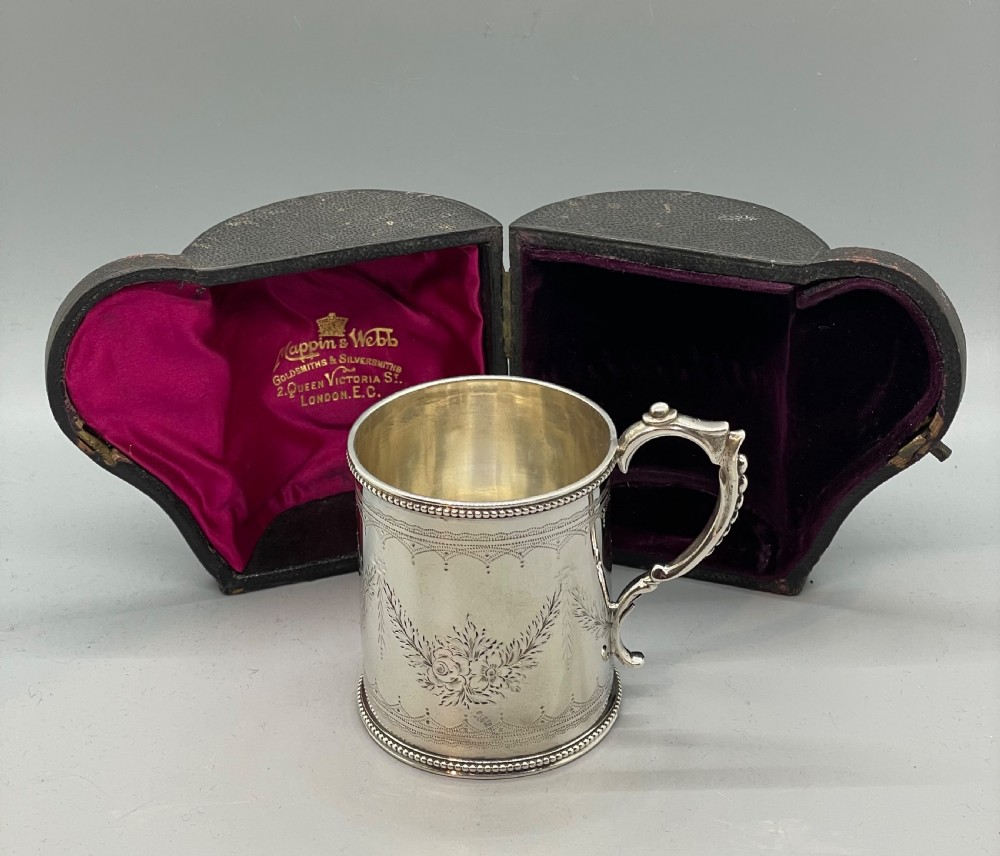 An antique sterling silver christening mug featuring floral engravings and beaded edges, placed beside its original fitted leather case lined with magenta velvet.