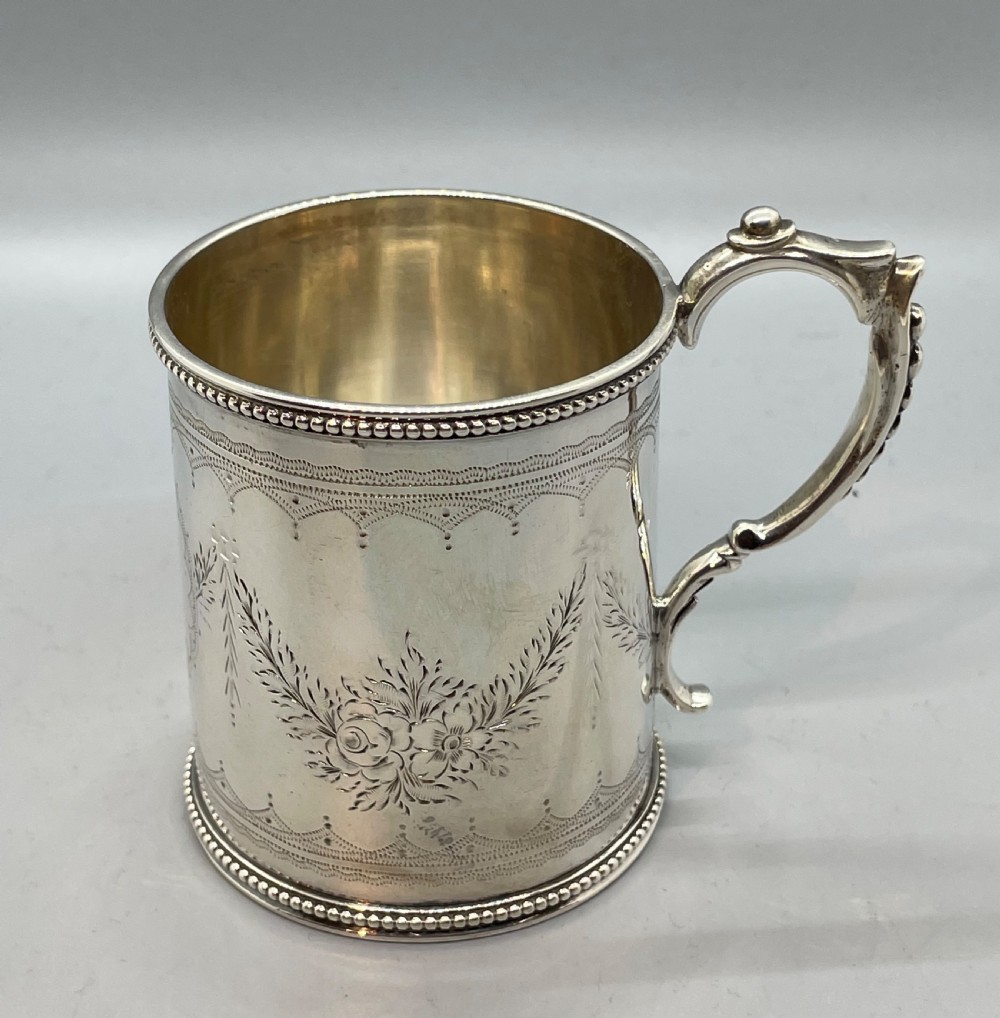An antique sterling silver christening mug featuring floral engravings and beaded edges, placed beside its original fitted leather case lined with magenta velvet. View II