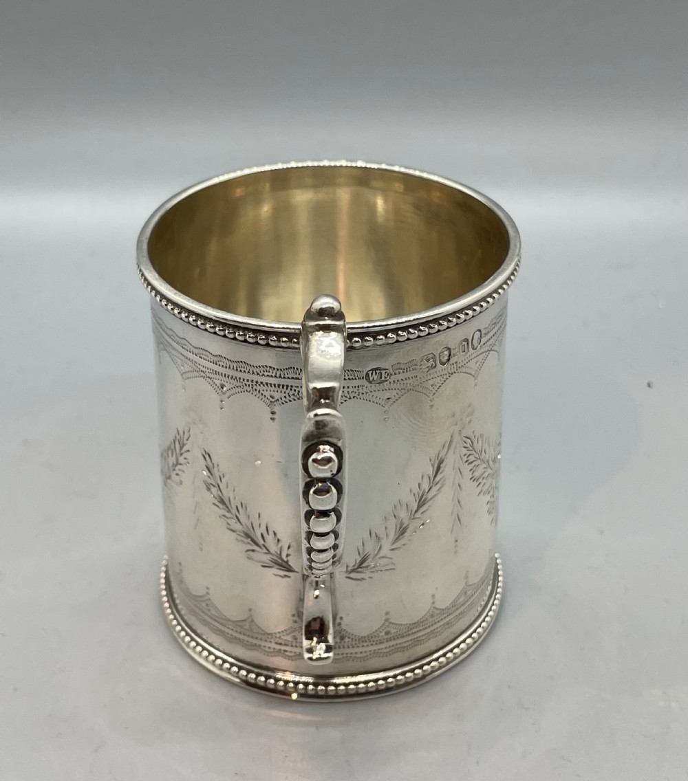 An antique sterling silver christening mug featuring floral engravings and beaded edges, placed beside its original fitted leather case lined with magenta velvet. View III