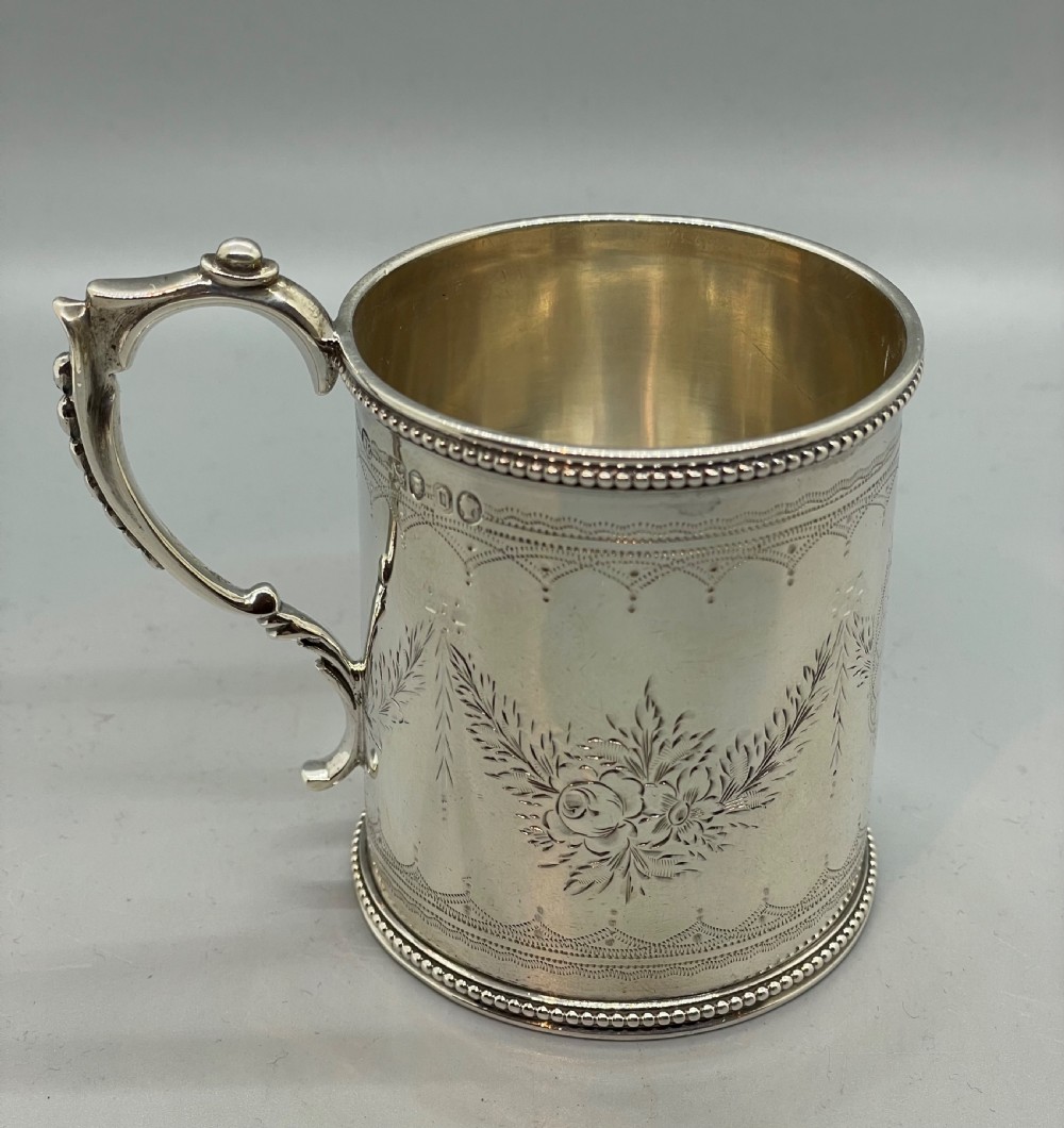 An antique sterling silver christening mug featuring floral engravings and beaded edges, placed beside its original fitted leather case lined with magenta velvet. View IV