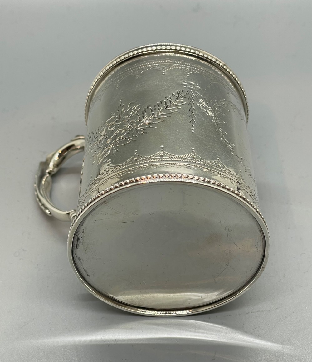 An antique sterling silver christening mug featuring floral engravings and beaded edges, placed beside its original fitted leather case lined with magenta velvet. View V