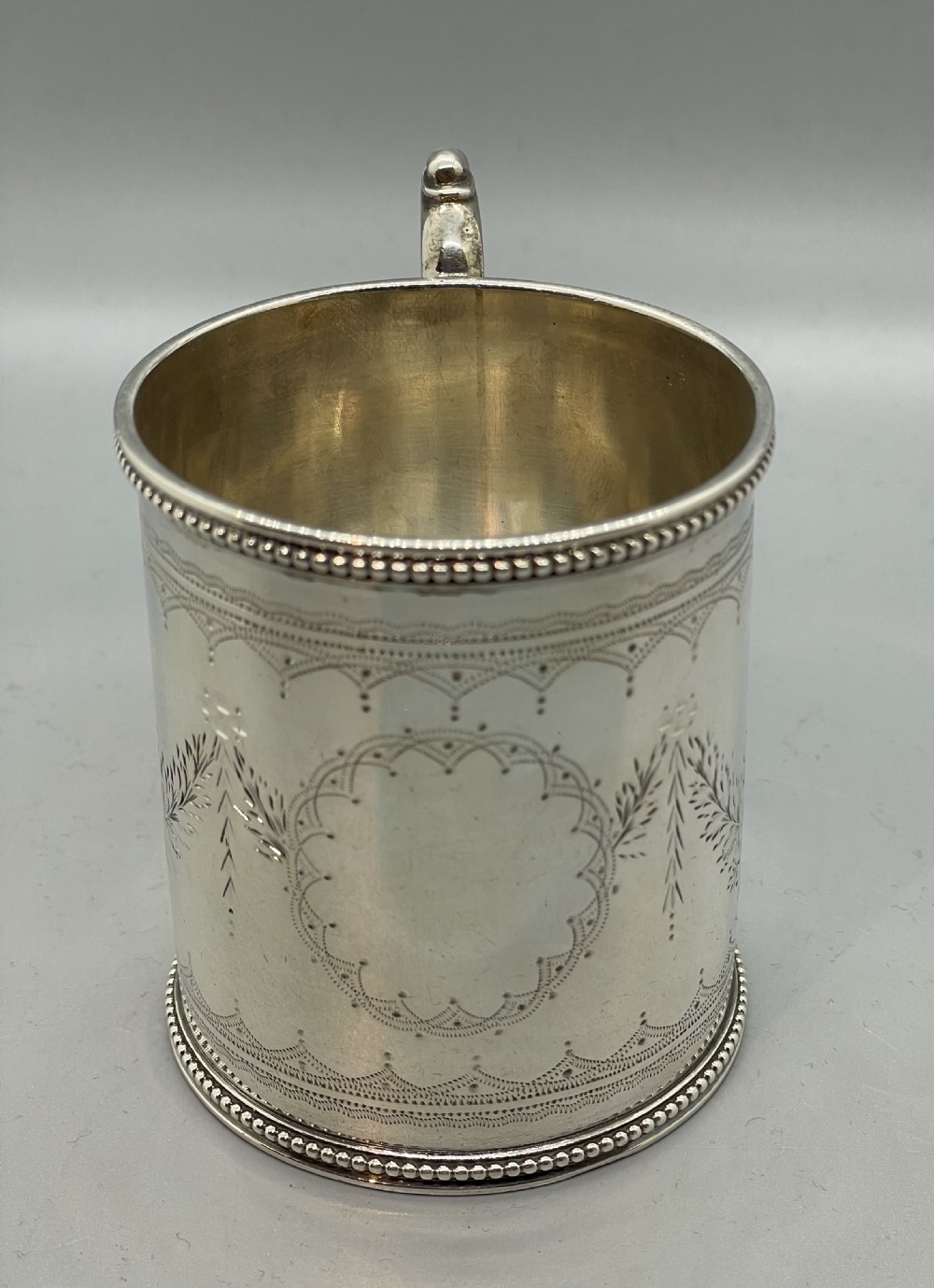 An antique sterling silver christening mug featuring floral engravings and beaded edges, placed beside its original fitted leather case lined with magenta velvet. View VI