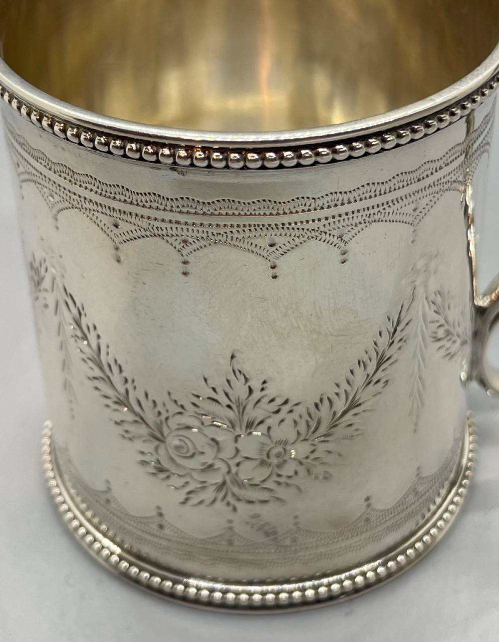 An antique sterling silver christening mug featuring floral engravings and beaded edges, placed beside its original fitted leather case lined with magenta velvet. View VIII
