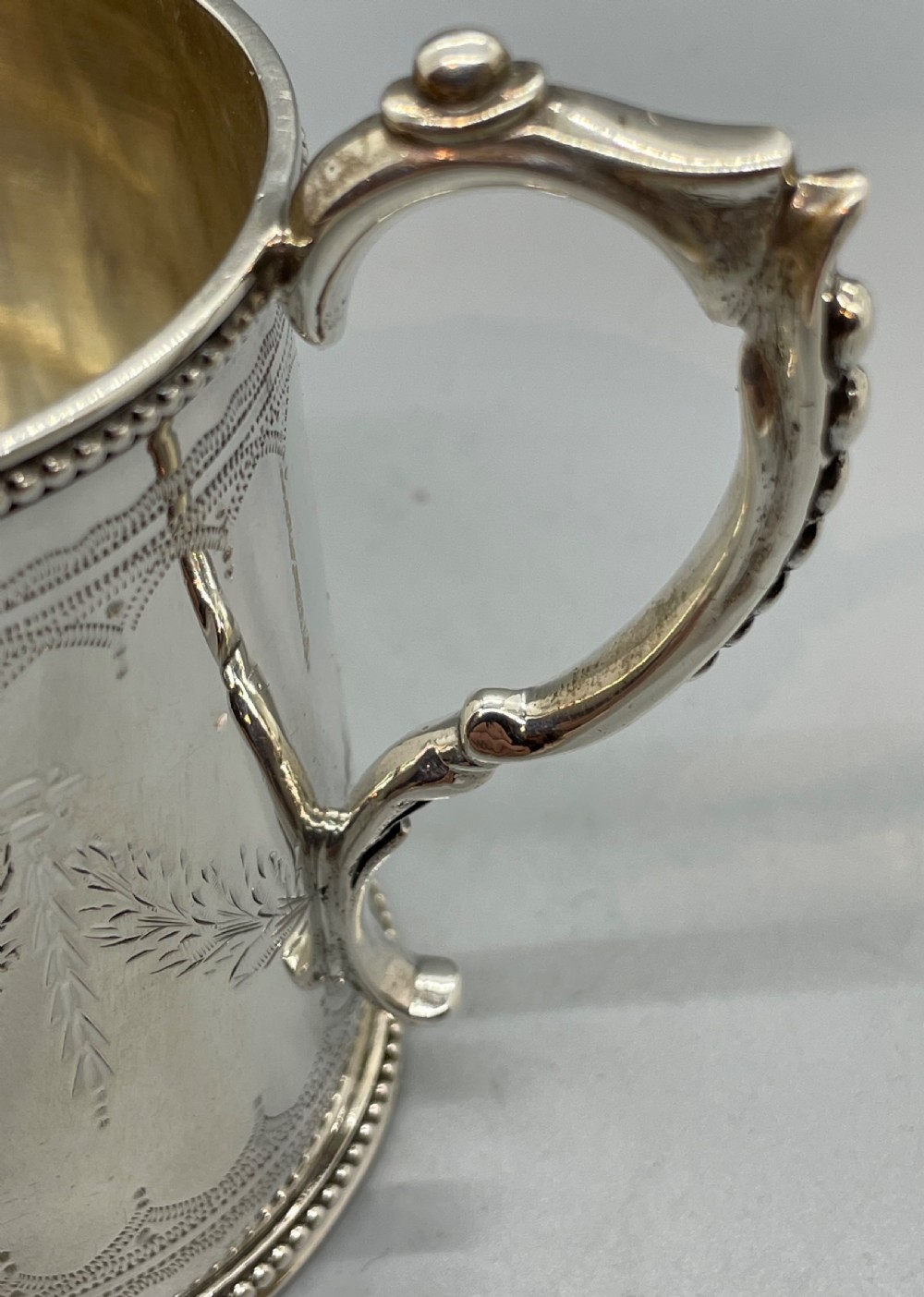 An antique sterling silver christening mug featuring floral engravings and beaded edges, placed beside its original fitted leather case lined with magenta velvet. View IX