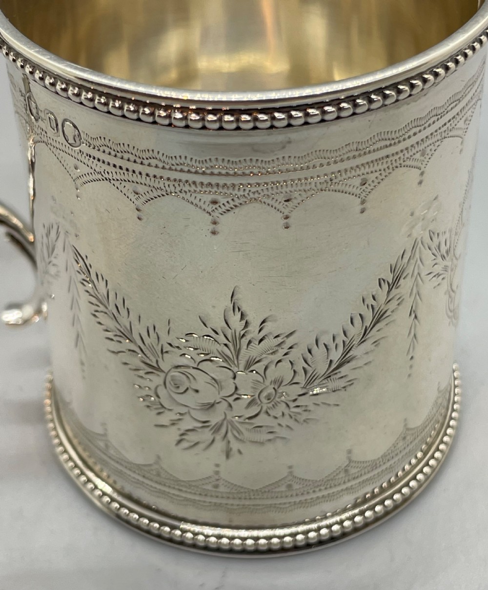 An antique sterling silver christening mug featuring floral engravings and beaded edges, placed beside its original fitted leather case lined with magenta velvet. View X