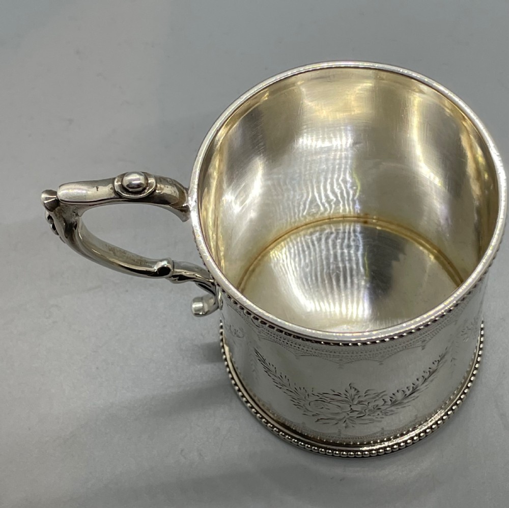 An antique sterling silver christening mug featuring floral engravings and beaded edges, placed beside its original fitted leather case lined with magenta velvet. View XI