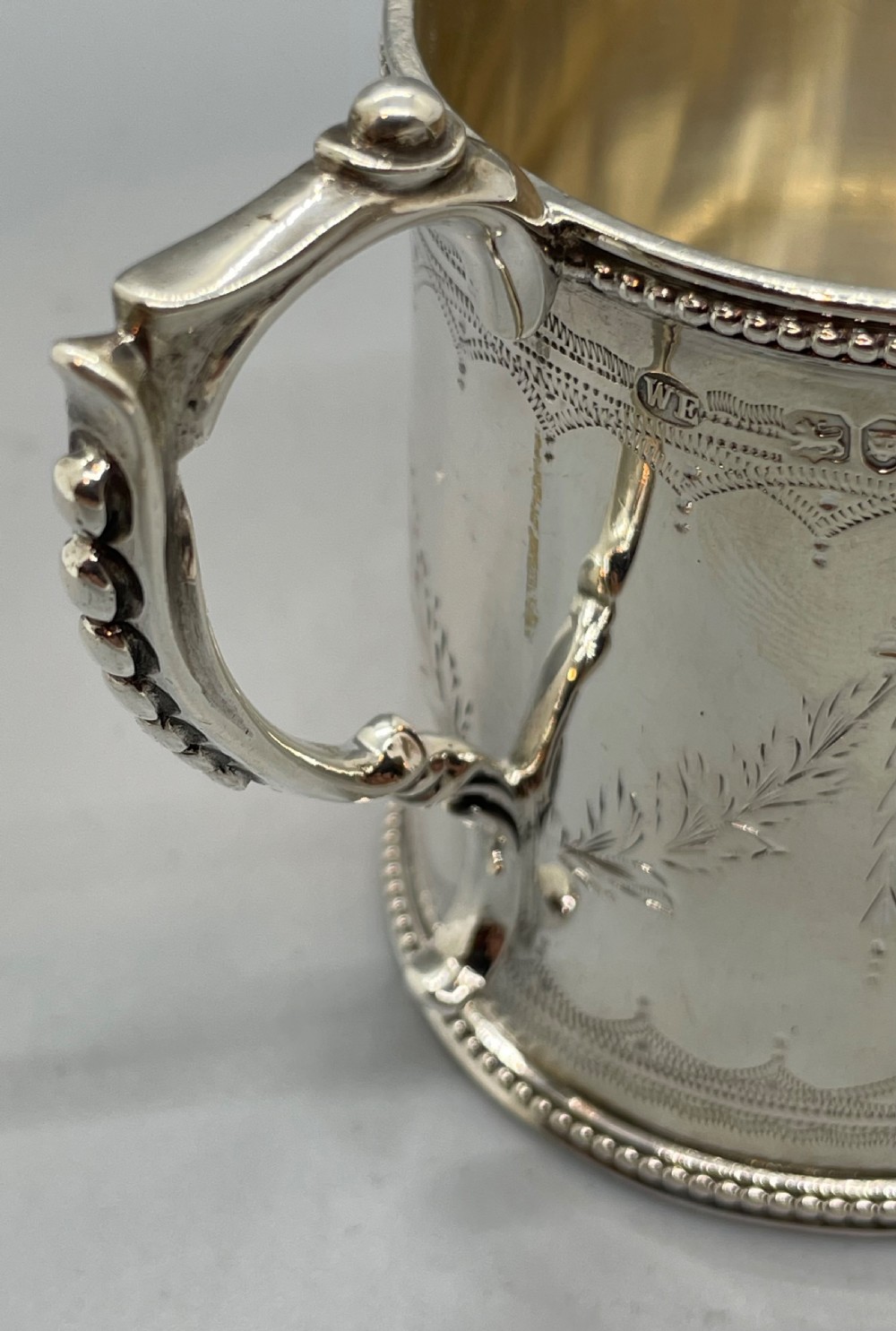 An antique sterling silver christening mug featuring floral engravings and beaded edges, placed beside its original fitted leather case lined with magenta velvet. View XIV