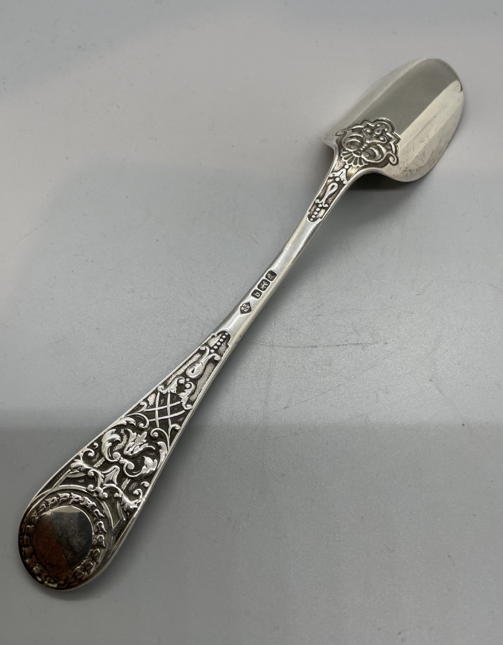 An Edwardian sterling silver stilton spoon displaying an ornate relief-carved handle with floral motifs and a polished scoop-shaped bowl. View V