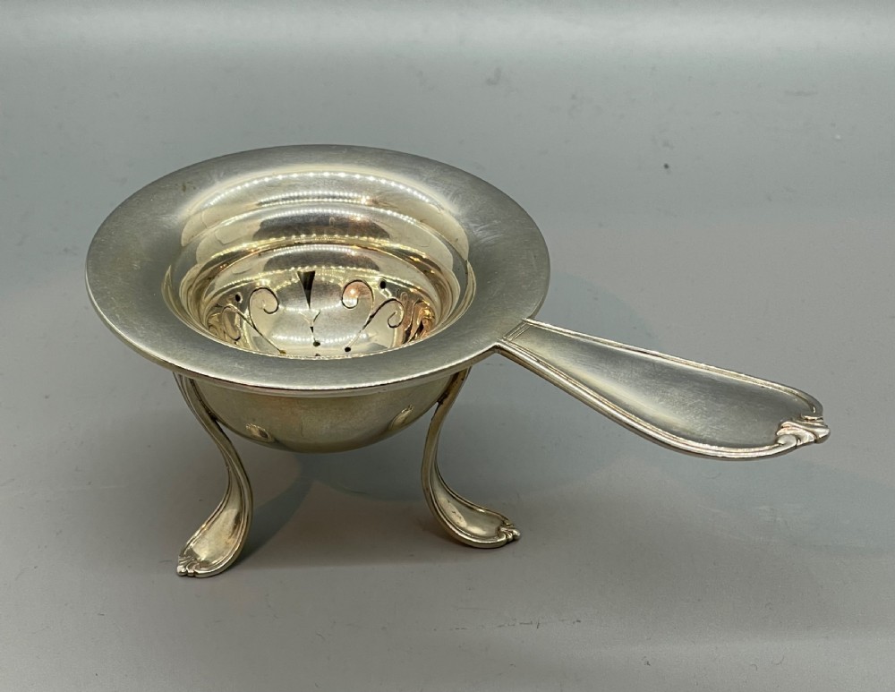 A Danish Art Deco silver tea strainer with a pierced bowl design and a smooth flat handle resting on a tripod stand.