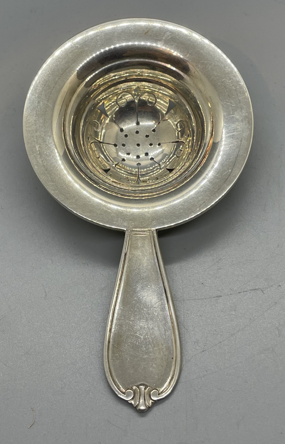 A Danish Art Deco silver tea strainer with a pierced bowl design and a smooth flat handle resting on a tripod stand. View VI