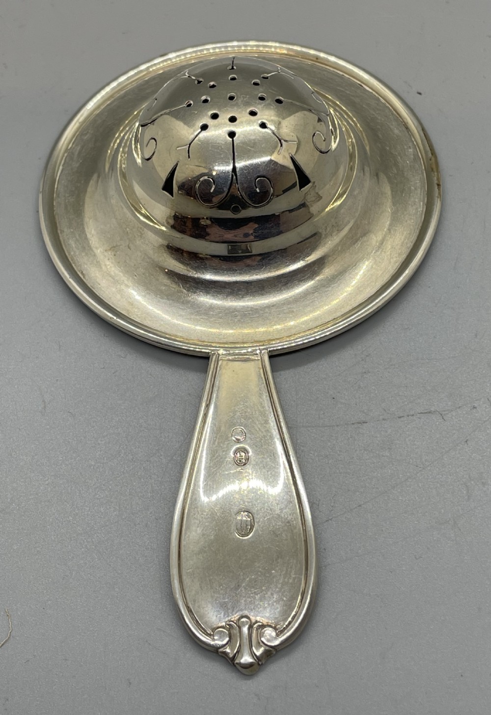A Danish Art Deco silver tea strainer with a pierced bowl design and a smooth flat handle resting on a tripod stand. View VII