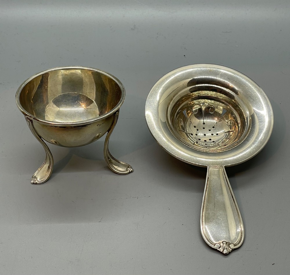 A Danish Art Deco silver tea strainer with a pierced bowl design and a smooth flat handle resting on a tripod stand. View X