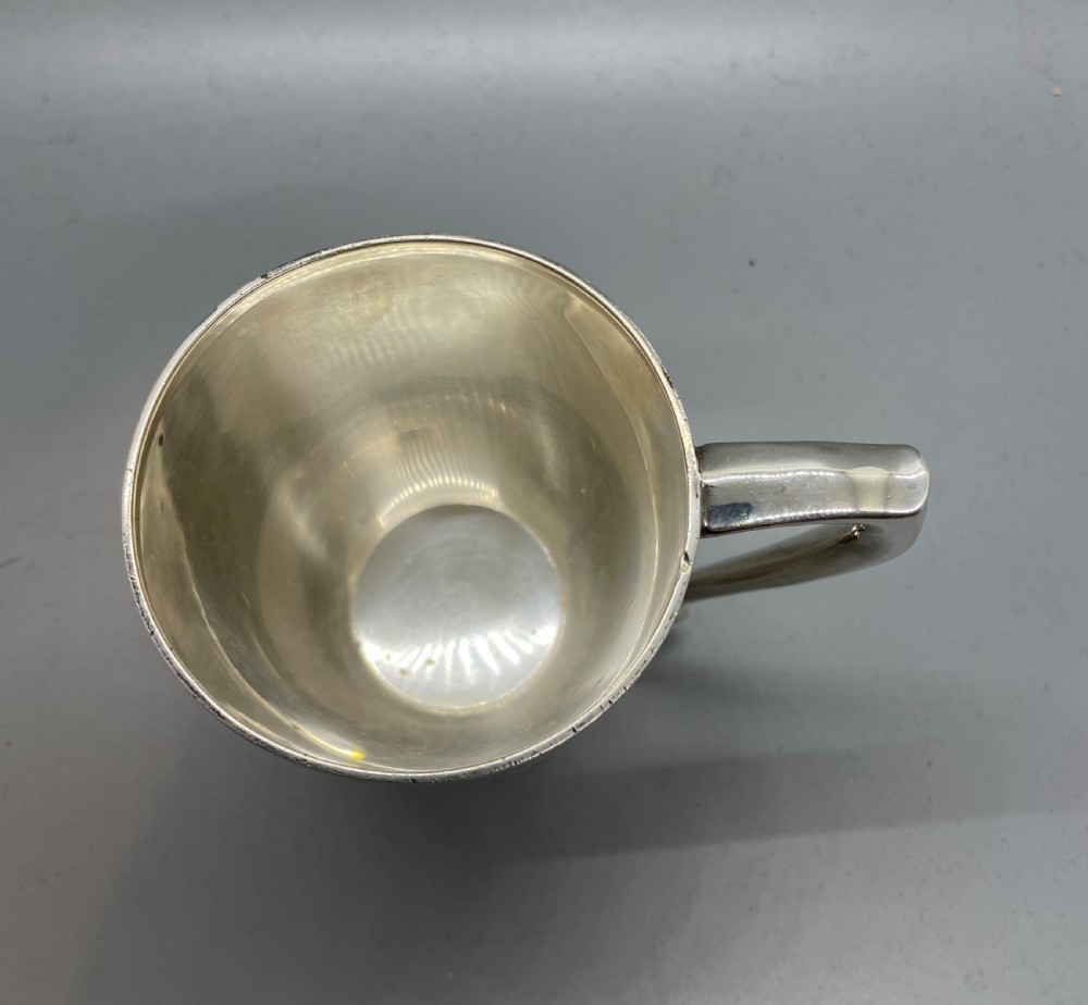 A polished sterling silver George V mug featuring a plain tapered body, an angular loop handle, and a scalloped pedestal base. View III