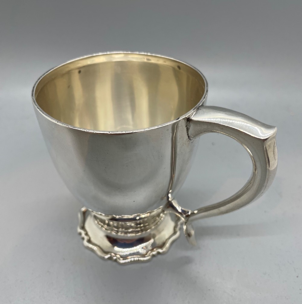 A polished sterling silver George V mug featuring a plain tapered body, an angular loop handle, and a scalloped pedestal base. View IV
