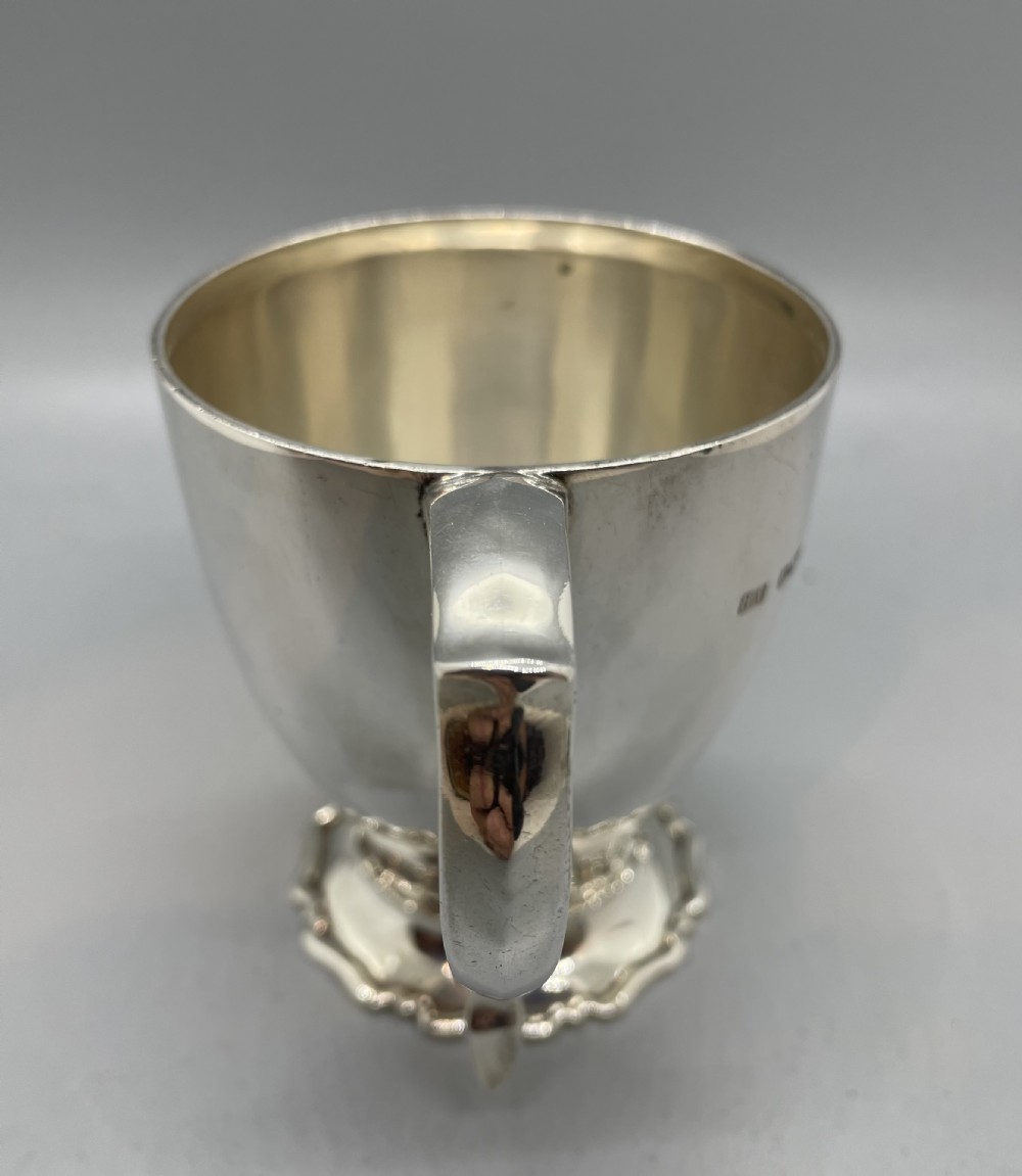 A polished sterling silver George V mug featuring a plain tapered body, an angular loop handle, and a scalloped pedestal base. View V
