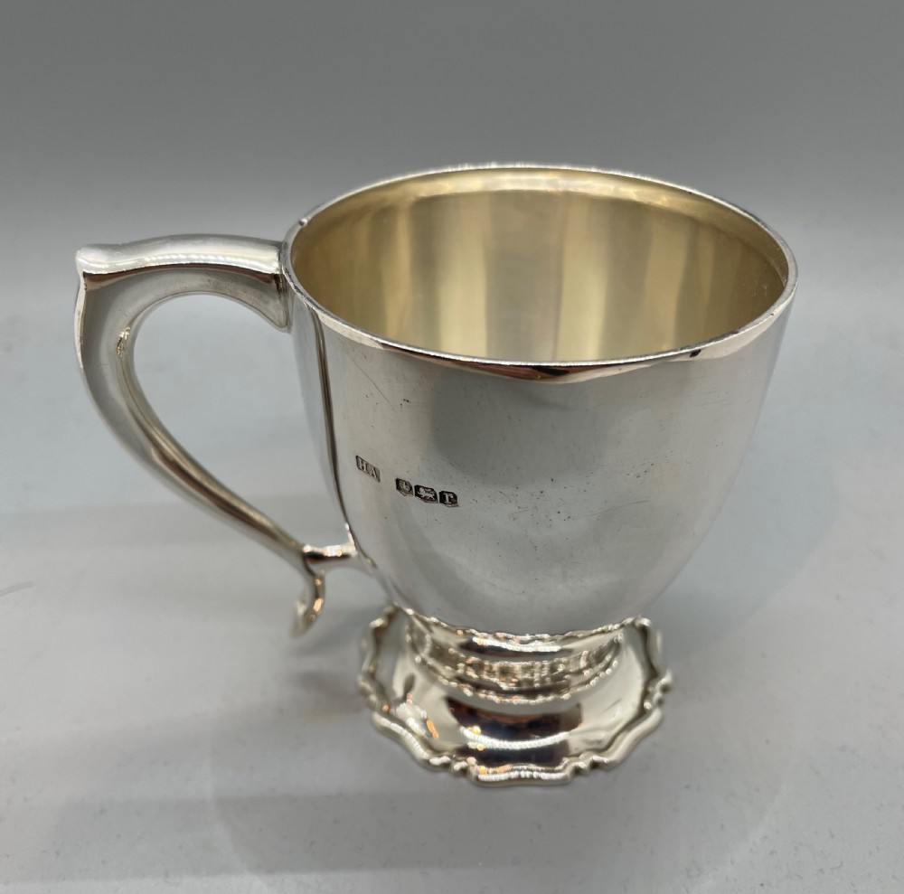 A polished sterling silver George V mug featuring a plain tapered body, an angular loop handle, and a scalloped pedestal base. View VI