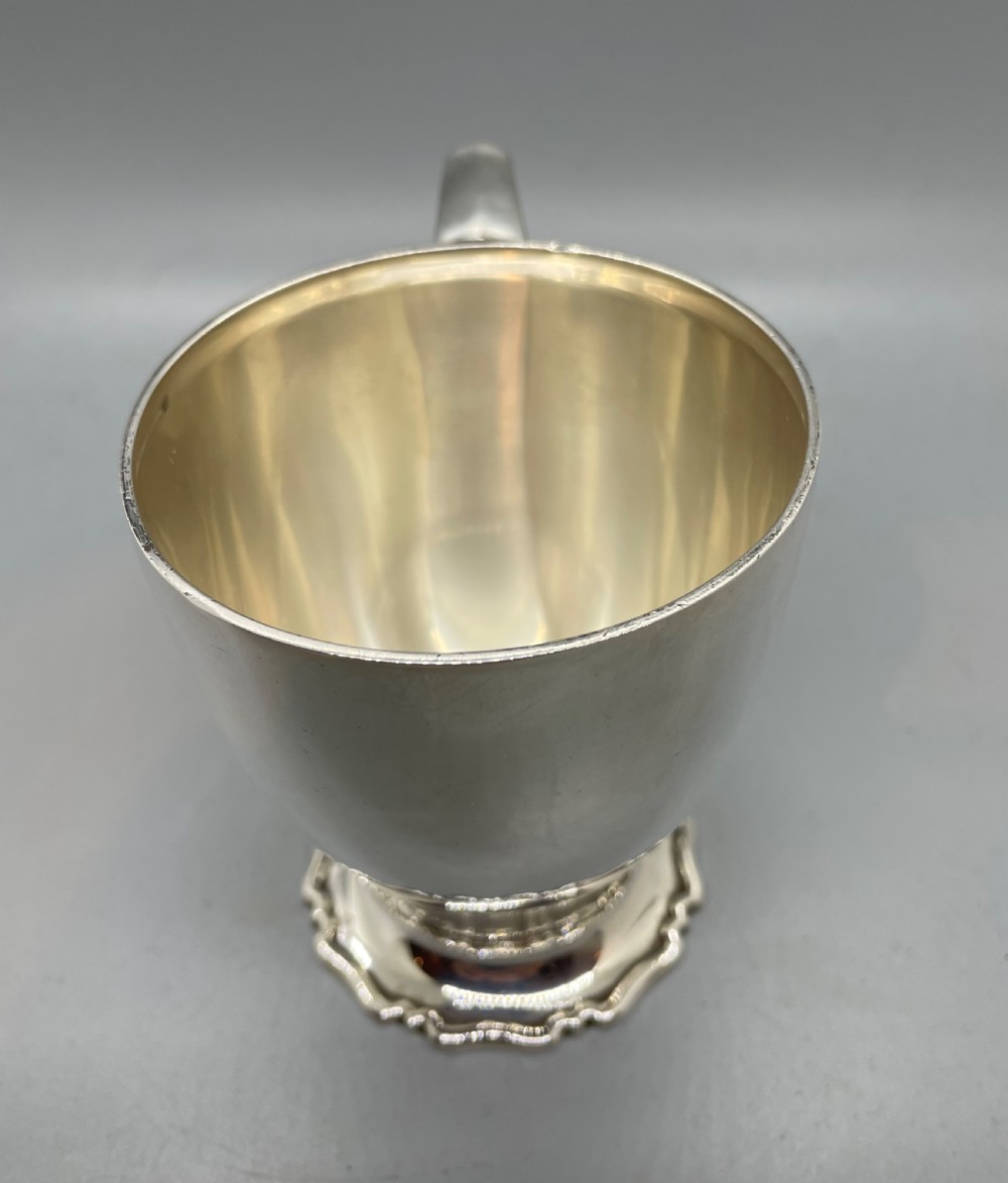 A polished sterling silver George V mug featuring a plain tapered body, an angular loop handle, and a scalloped pedestal base. View VII