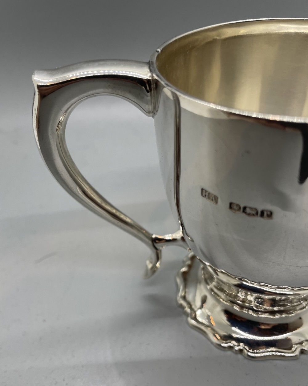 A polished sterling silver George V mug featuring a plain tapered body, an angular loop handle, and a scalloped pedestal base. View VIII