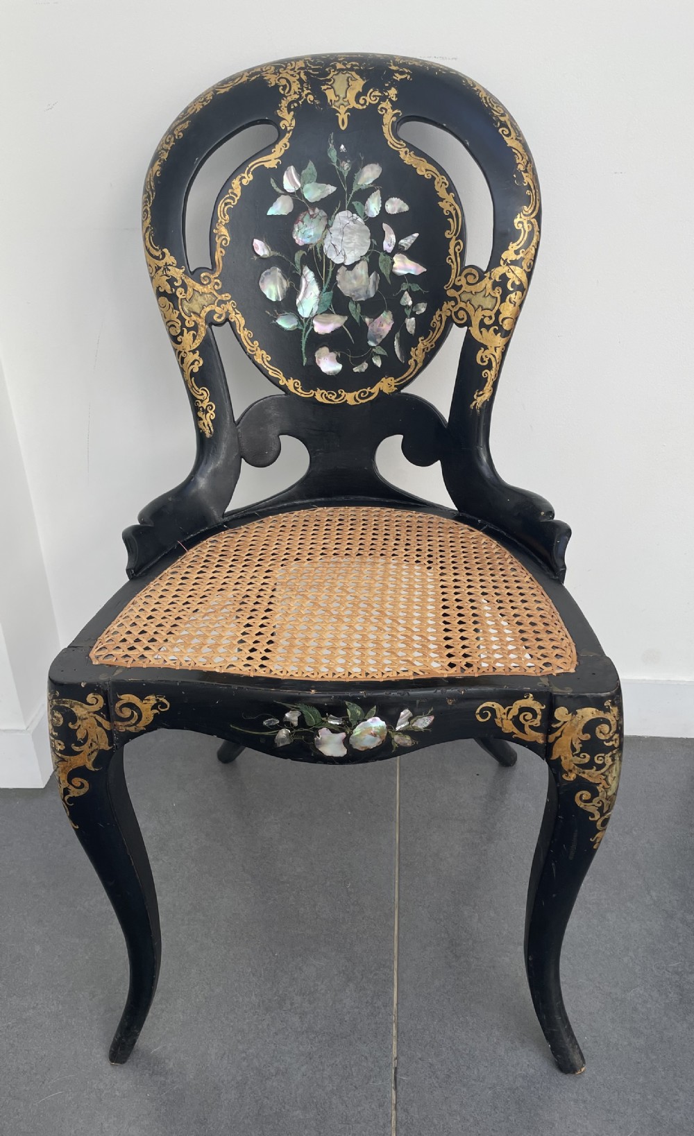 A Victorian ebonised side chair featuring floral mother-of-pearl inlays, gold leaf scrollwork decoration, a shaped back rest, cabriole front legs, and a woven cane seat.