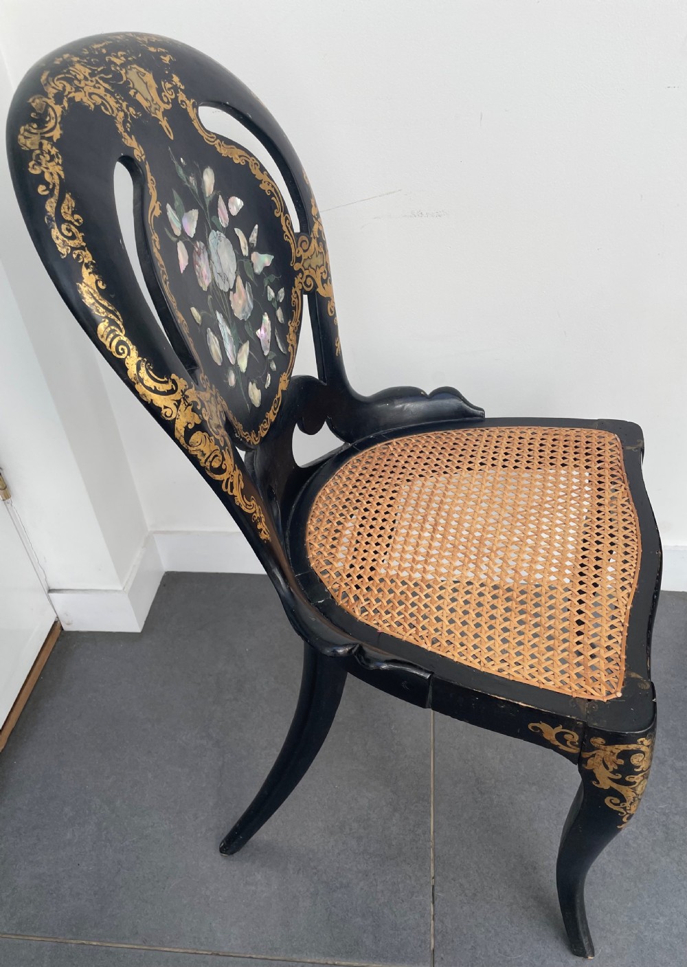 A Victorian ebonised side chair featuring floral mother-of-pearl inlays, gold leaf scrollwork decoration, a shaped back rest, cabriole front legs, and a woven cane seat. View VI