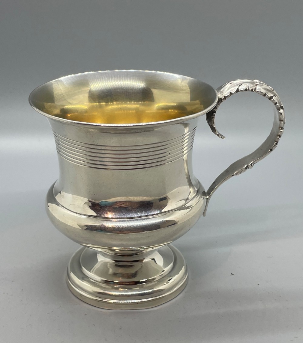 A George IV sterling silver mug featuring horizontal reeded decoration, a leaf-scrolled handle, a molded pedestal foot, and a gold-gilded interior.